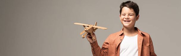 panoramic shot of amused boy holding wooden toy plane isolated on grey - Foto, Imagem