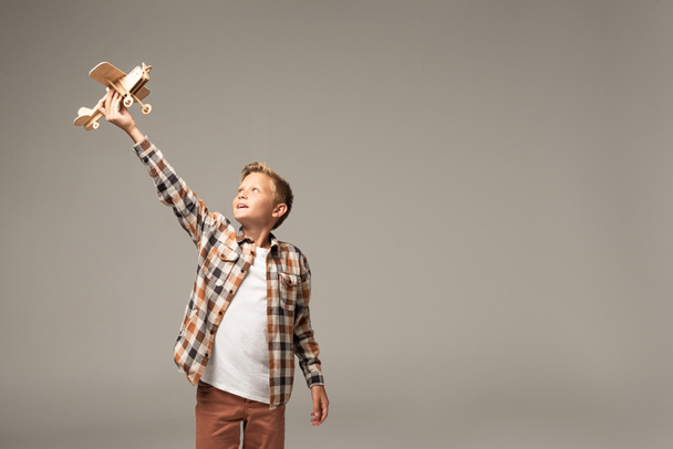 happy boy holding wooden toy plane in raised hand isolated on grey - Foto, Imagem