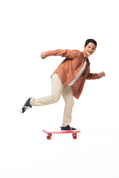 cheerful boy riding penny board while looking at camera on white background - Photo, image
