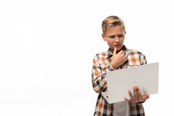 przemyślany chłopiec patrząc na laptopa i trzymając się za rękę blisko twarzy odizolowany na białym - Zdjęcie, obraz