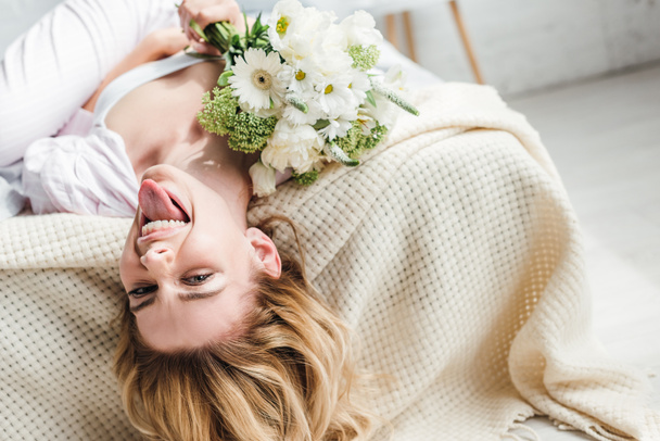 selective focus of happy girl sticking out tongue while lying on bed with flowers  - Fotoğraf, Görsel