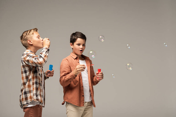 boy blowing soap bubbles near amazed brother isolated on grey - Fotografie, Obrázek
