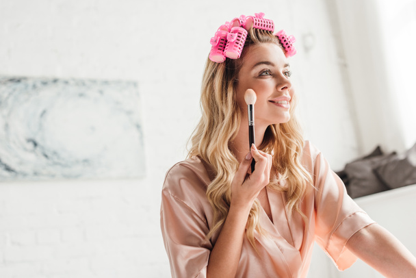 veselá žena v růžové vlasy natáčky drží kosmetické štětec v blízkosti obličeje v ložnici  - Fotografie, Obrázek