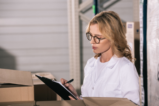 concentrated storekeeper in glasses writing on clipboard in warehouse - Photo, Image