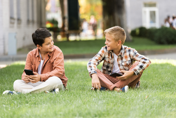dva usměvaví bratři mluví, zatímco sedí na trávníku s chytrými telefony - Fotografie, Obrázek