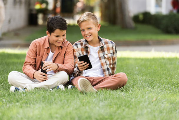 two smiling brothers sitting on green grass and using smartphones - Fotoğraf, Görsel