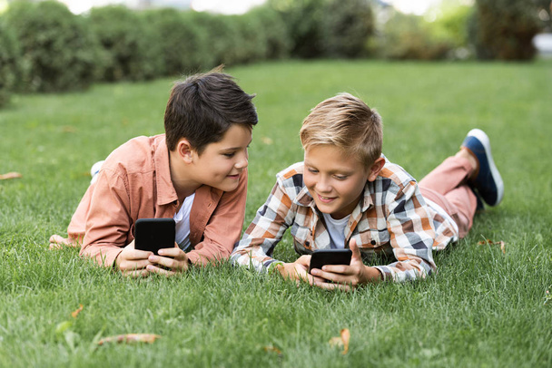 two cheerful brothers lying on green lawn and using smartphones - 写真・画像