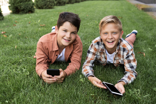 two cheerful brothers lying on grass, holding smartphones and looking at camera - Fotoğraf, Görsel