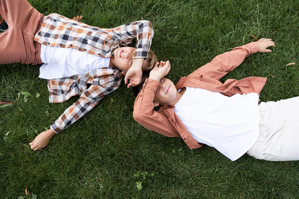 top view of two smiling brothers covering eyes with hands while lying on grass - Fotografie, Obrázek