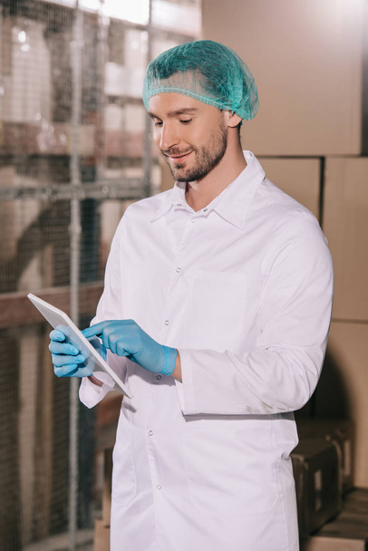 smiling storekeeper in white coat and hairnet using digital tablet in warehouse - Foto, Imagen
