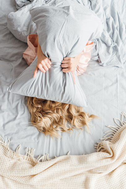 vista superior de la joven mujer cubriendo la cara con almohada y acostado en la cama
  - Foto, Imagen
