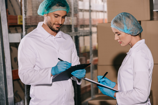 two storekeepers in white coats and hairnets writing on clipboards in warehouse - Foto, Imagen
