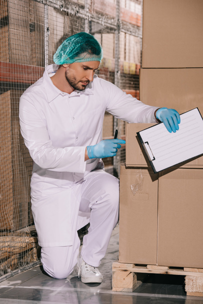 almacenista concentrado inspeccionando la caja de cartón mientras sostiene el portapapeles
 - Foto, Imagen
