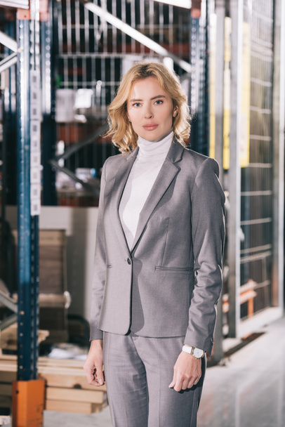 confident businesswoman in formal wear looking at camera while standing in warehouse - Фото, изображение