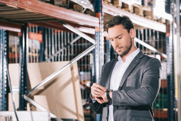 serious businessman checking time on wristwatch while standing in warehouse - 写真・画像