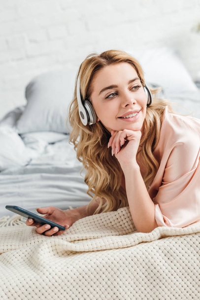 happy girl in headphones listening music and holding smartphone in bedroom  - Fotografie, Obrázek