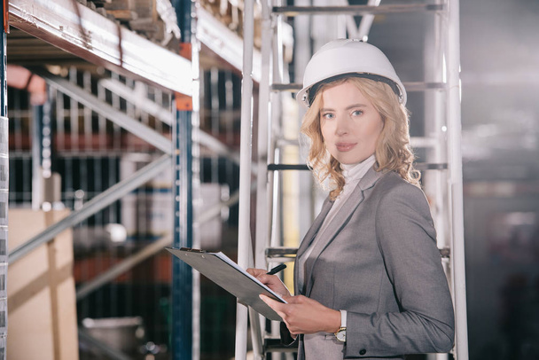confident businesswoman in helmet holding clipboard and looking at camera - Photo, Image