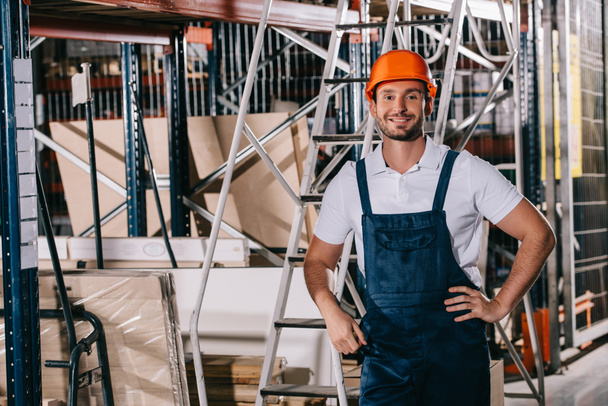 handsome warehouse worker standing with hands on hips and smiling at camera - Photo, Image
