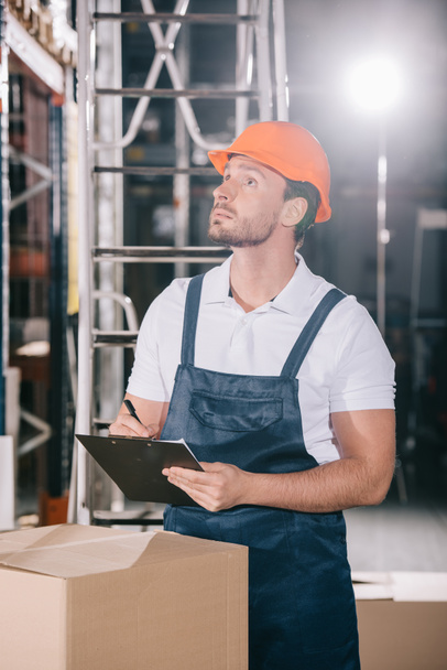 concentrated warehouse worker looking away while writing on clipboard - Photo, Image