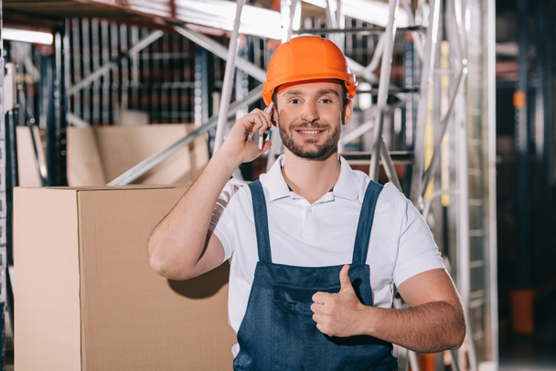 smiling warehouse worker talking on smartphone, looking at camera and showing thumb up - 写真・画像