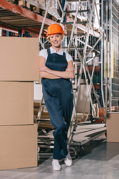 trabajadora sonriente mirando a la cámara mientras está de pie cerca de cajas de cartón con brazos cruzados
 - Foto, imagen