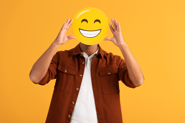 KYIV, UKRAINE - SEPTEMBER 24, 2019: man covering face with happy emoticon isolated on orange  - Photo, image