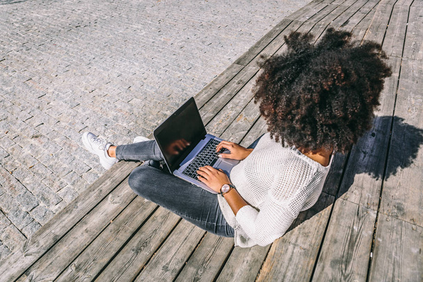 Chica morena con pelo afro y ropa casual trabaja con su portátil en un área al aire libre
 - Foto, Imagen