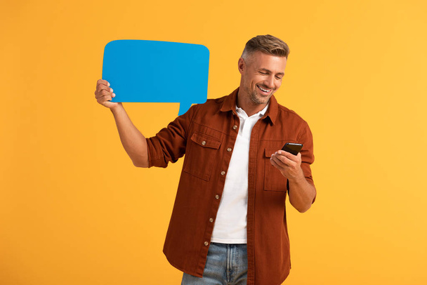 happy man holding blue speech bubble and smartphone isolated on orange  - Foto, immagini