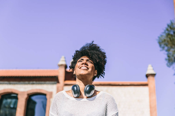 Beautiful dark skinned woman with big black headphones around her neck, smiles looking up while walking through - Fotó, kép