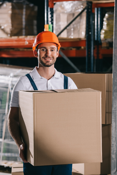 cheerful loader smiling at camera while holding cardboard box - Foto, afbeelding