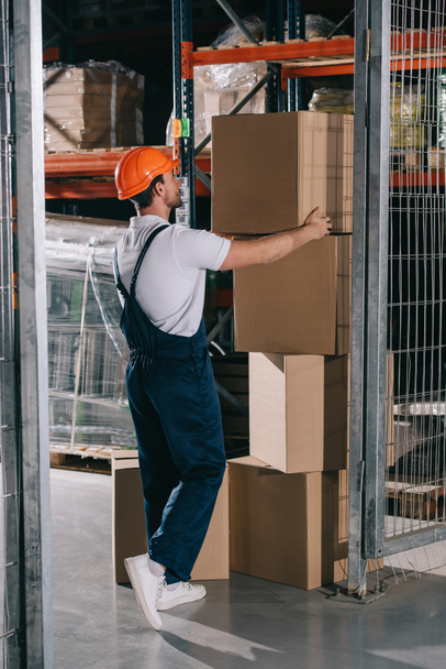 работник склада в комбинезоне и шлеме берет картонную коробку из стопки
 - Фото, изображение