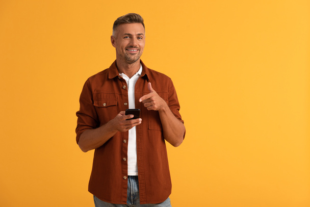 happy man pointing with finger at smartphone isolated on orange  - Foto, Bild