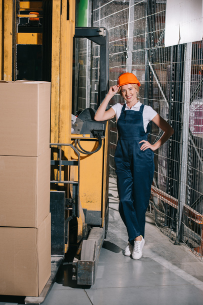 alegre trabajadora de pie cerca de la carretilla elevadora con la mano en la cadera, tocando el casco y mirando a la cámara
 - Foto, Imagen