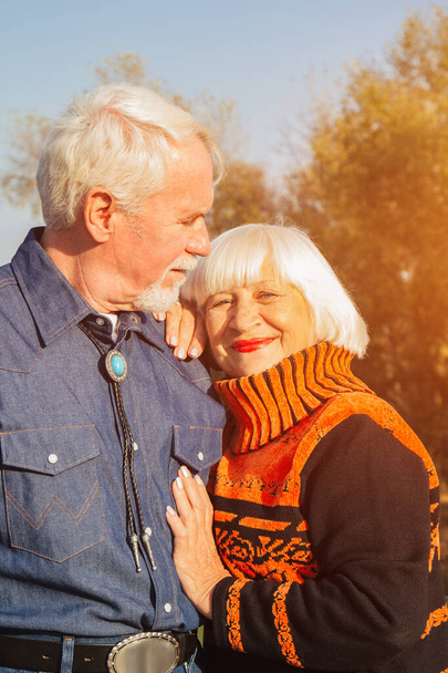 Happy elderly man and woman sitting on a bench in autumn day. Relaxed senior couple sitting on a park bench. Grandfather gently kisses grandmother on the forehead. - Foto, immagini