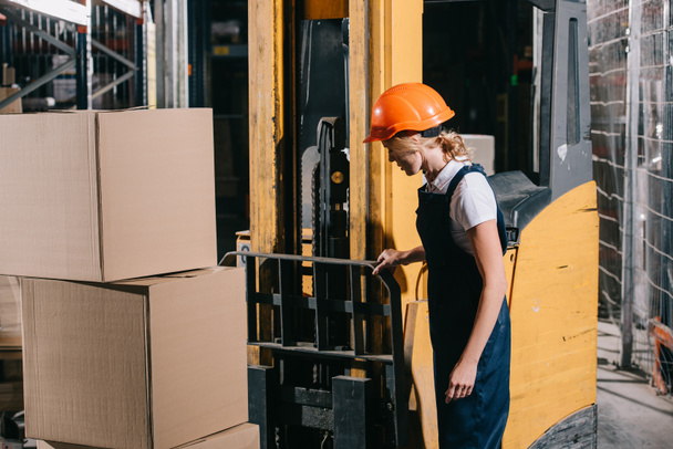 trabajadora en overol y casco de pie cerca del cargador de foklift en el almacén
 - Foto, Imagen
