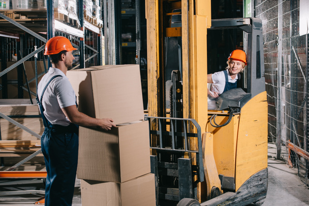 lachende werkman zittend in vorkheftruck lader en kijkend naar werknemer die kartonnen doos vasthoudt - Foto, afbeelding