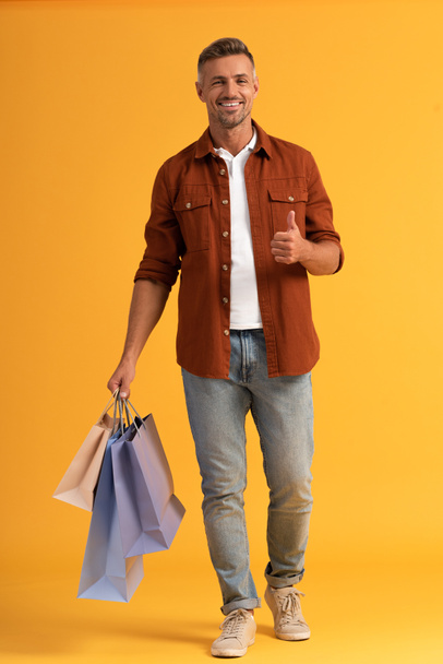 cheerful man holding colorful shopping bags and showing thumb up on orange  - Valokuva, kuva