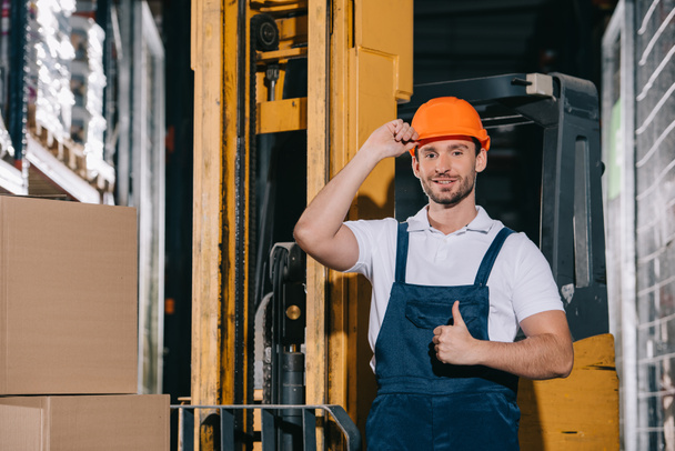 smiling warehouse worker touching helmet and showing thumb up while standing near forklift loader - Φωτογραφία, εικόνα