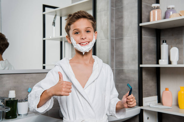 χαρούμενο αγόρι με αφρό ξυρίσματος στο πρόσωπο κρατώντας ξυράφι και δείχνοντας τον αντίχειρα επάνω  - Φωτογραφία, εικόνα
