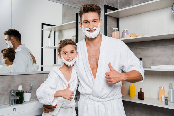 cheerful father and son with shaving foam on faces showing thumbs up in bathroom  - Fotó, kép