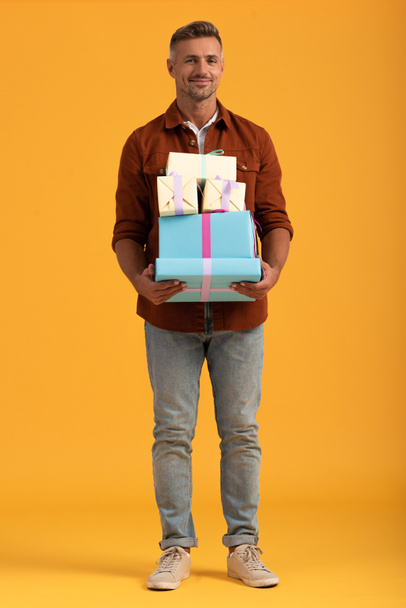 handsome man smiling while holding gift boxes on orange  - Foto, afbeelding