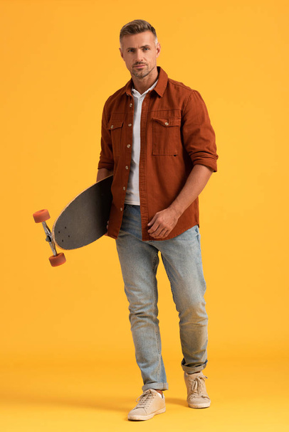 handsome man standing and holding penny board on orange - Zdjęcie, obraz