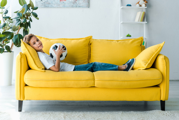 glücklicher Junge im weißen T-Shirt auf gelbem Sofa liegend mit Fußball  - Foto, Bild