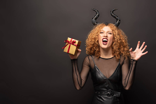 beautiful redhead woman in halloween costume with horns holding gift box on black - Фото, изображение