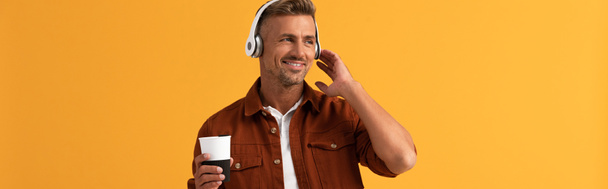 panoramisch schot van gelukkige man luisteren muziek en het houden van koffie te gaan geïsoleerd op oranje  - Foto, afbeelding