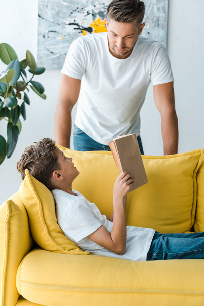 happy and cute kid holding book while lying on sofa and looking at father at home  - Photo, Image