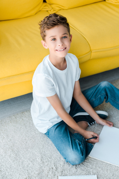 glückliches Kind sitzt auf Teppich neben gelbem Sofa zu Hause  - Foto, Bild