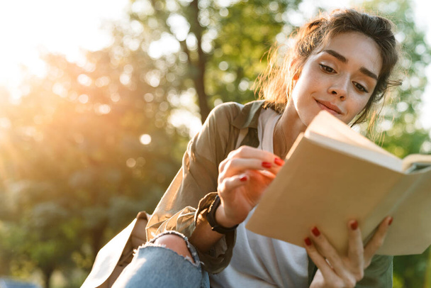 Image of young woman smiling and reading book in green park - Photo, image