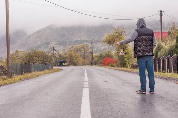 Φθινοπωρινό πρωινό στο βουνό, ένας τύπος με την πλάτη του να στέκεται στο δρόμο περιμένοντας ένα διερχόμενο αυτοκίνητο να φτάσει εκεί. - Φωτογραφία, εικόνα
