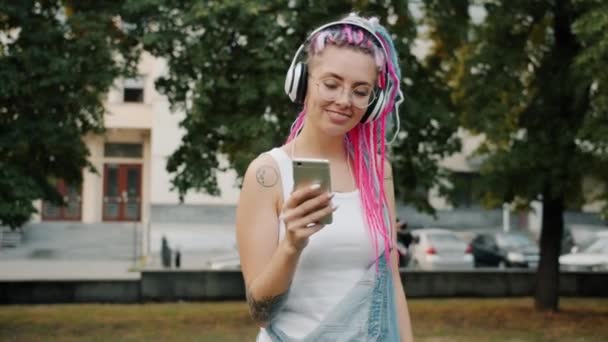 Happy hipster in headphones dancing outdoors in park using smartphone - Felvétel, videó